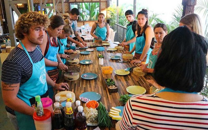 Tourists in a Thai cooking class learn to prepare traditional dishes, gathered around a table with fresh ingredients and utensils