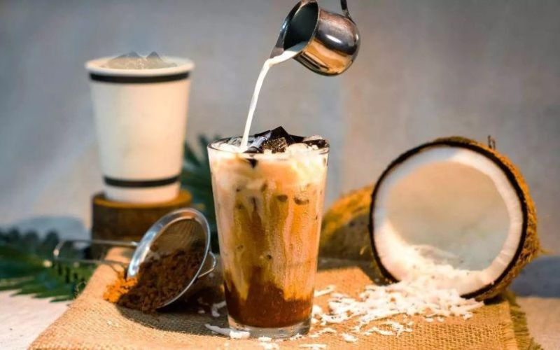 Iced coconut coffee being poured with coconut milk, served in a tall glass beside fresh coconut shells and shredded coconut