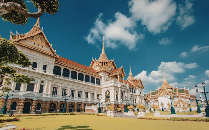 A wide-angle landscape shot of the grand Chakri Maha Prasat Throne Hall in the Grand Palace, with its European-inspired architecture and three traditional Thai spires, surrounded by manicured lawns and pruned trees under a blue sky.