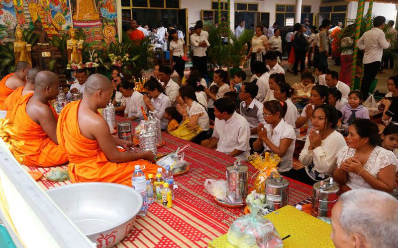 Buddhist monks in orange robes receiving offerings from families seated on mats during Pchum Ben ancestor festival in Cambodia.