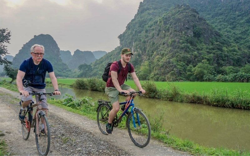 Two travelers riding bicycles along a rural path beside a small canal, surrounded by lush green rice fields and limestone mountains in Vietnam’s countryside.