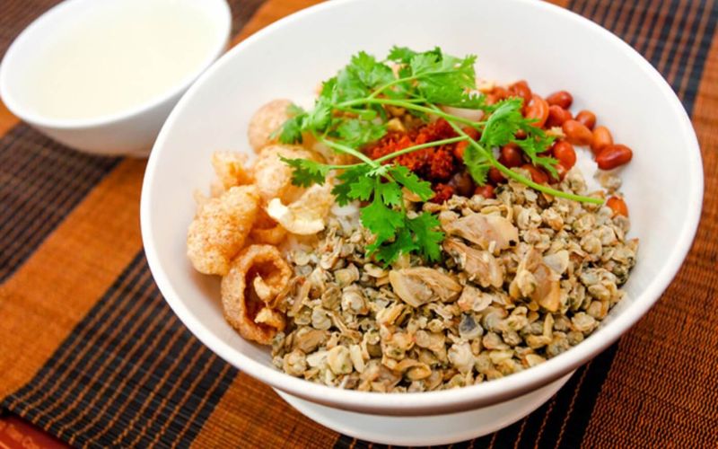 A bowl of Com Hen, a traditional Hue clam rice dish, featuring a base of white rice topped with a generous portion of small stir-fried clams, crispy pork rinds, roasted peanuts, chili paste, and fresh cilantro, served with a small bowl of clam broth on the side.