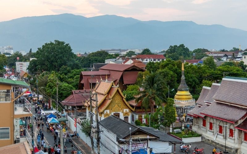 An elevated view looking over the Old City of Chiang Mai at dusk. The scene shows a blend of traditional Thai temple architecture (including a golden chedi with a tiered roof and orange-tiled temples) nestled among dense trees, bordering a busy local walking street or night market. Rolling blue mountains (Doi Suthep) form the backdrop under a soft evening sky.
