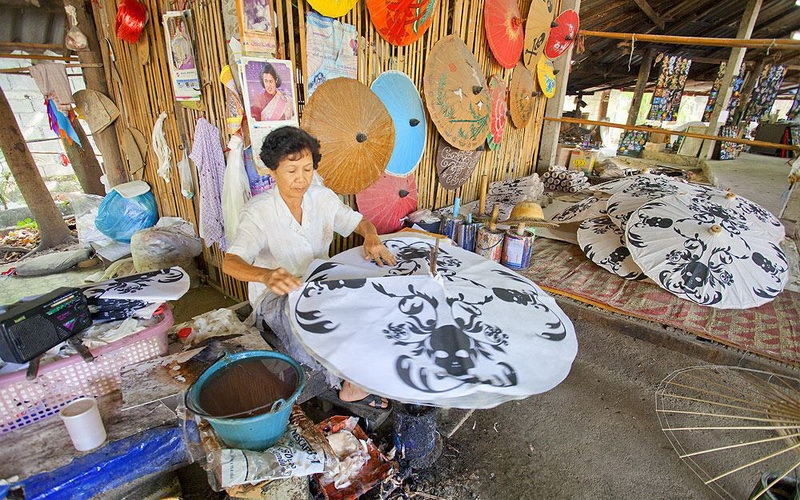 A close-up photograph of a woman wearing a white shirt hand-painting black patterns, including skulls, onto a large white umbrella/parasol in a rustic, open-air workshop at the Bo Sang and Sankhampaeng craft village.