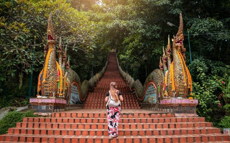 A lone female traveler, seen from behind, stands at the bottom of the grand, steep red-tiled staircase leading up to Wat Phra That Doi Suthep in Chiang Mai, Thailand. The staircase is flanked on both sides by elaborate, multi-headed Nāga (serpent) sculptures adorned with golden scales and floral garlands, extending far into the lush green forest at the top. Sunlight filters through the trees at the top of the stairs.