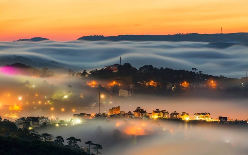 Aerial view of Da Lat city covered in sunset mist, with pine-covered hills, rooftops peeking through the fog, and soft golden sunlight filtering in from the horizon.