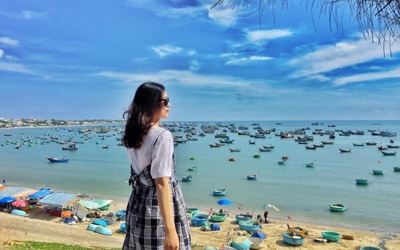 A woman stands on a coastal hill overlooking a sea full of fishing boats under a bright blue sky in southern Vietnam during the dry season.