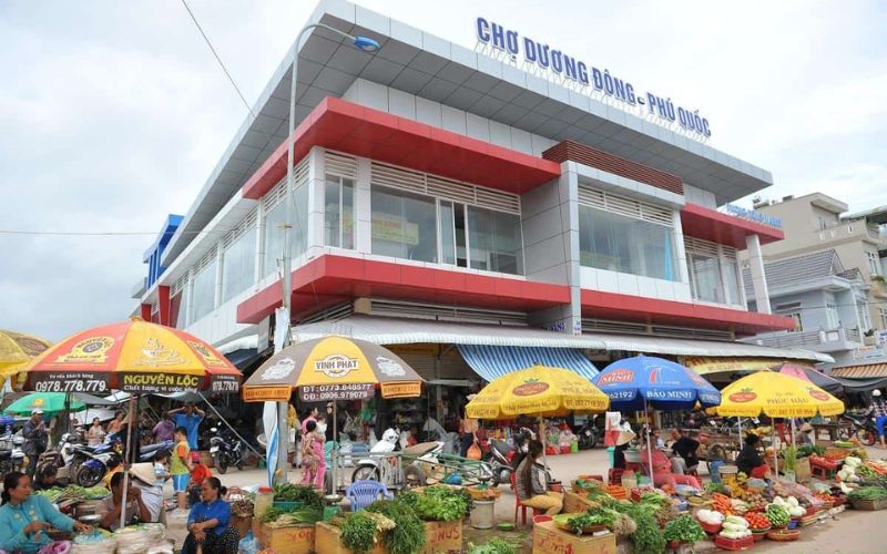 A bustling street-level view of the modern two-story Duong Dong Market building in Phu Quoc, with its grey and red facade and numerous colorful umbrellas covering outdoor vendors selling fresh produce.