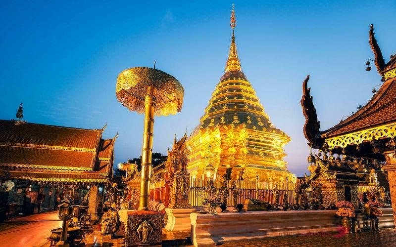 A brightly illuminated golden stupa at Doi Suthep Temple in Chiang Mai, surrounded by ornate structures, statues, and decorative umbrellas under a clear blue evening sky.