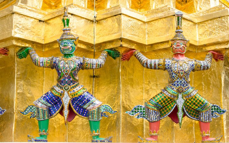 A close-up view of two colorful Yaksa (guardian giant) statues in traditional, ornate ceremonial attire, flanking and supporting the base of a gilded golden structure at Wat Phra Kaew.