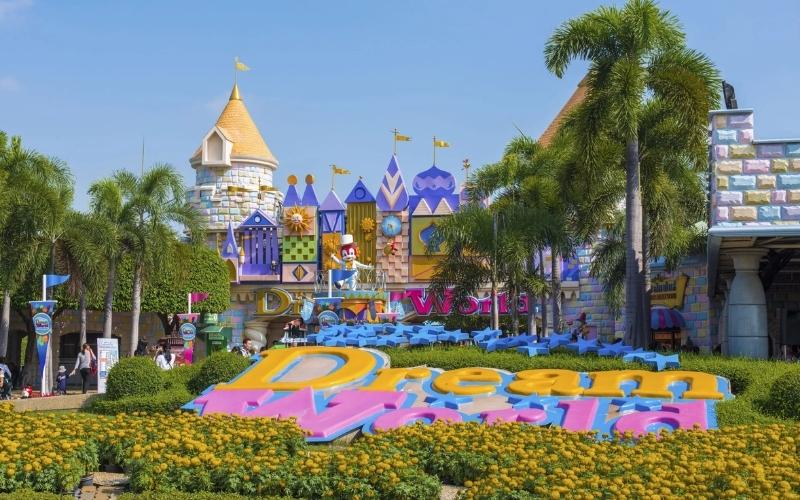 The whimsical, castle-like entrance to Dream World Bangkok amusement park, featuring colorful facades and a large 3D "Dream World" sign surrounded by yellow flowers and palm trees.