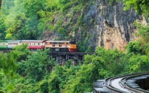 A train travels along the historic Death Railway in Kanchanaburi, Thailand, passing through lush greenery and rugged cliffside tracks
