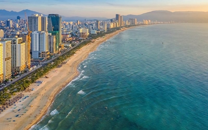 Aerial view of a long sandy beach bordered by turquoise waters and a skyline of modern high-rise buildings under a warm sunset in a coastal city.