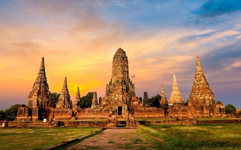 A breathtaking sunset view of the ancient brick pagodas and central prang at Wat Chaiwatthanaram in Ayutthaya Historical Park, with the sky painted in shades of orange and purple.