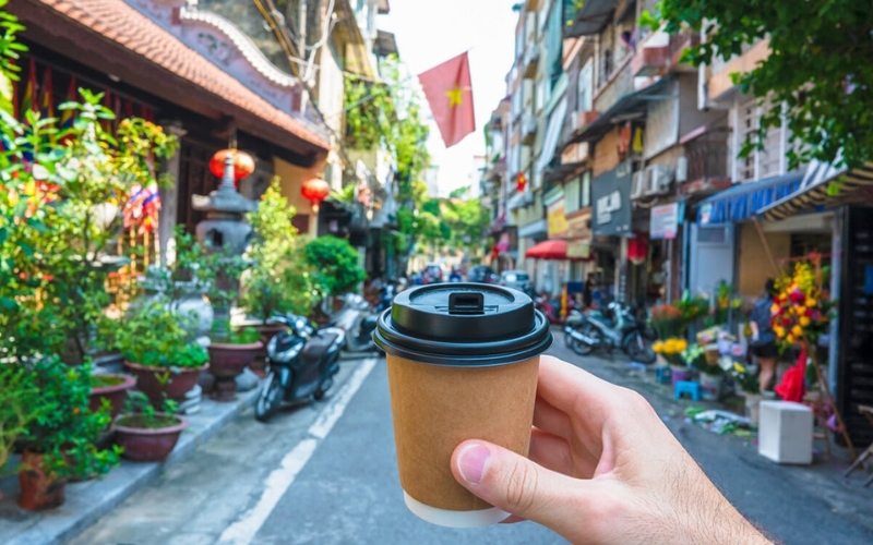 A traveler holds a fresh cup of Vietnamese coffee while strolling through a lively street in Hanoi’s Old Quarter, a perfect backdrop for experiencing the city’s rich coffee culture.