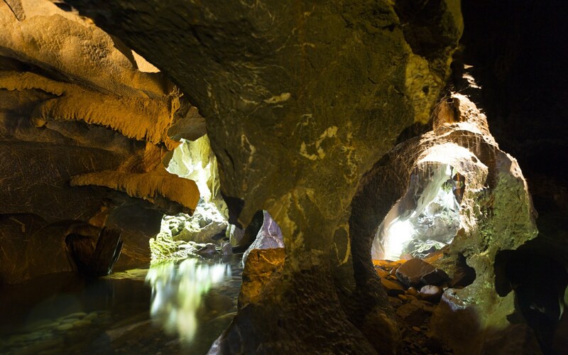 Inside Nam Thalu Cave in Khao Sok National Park, featuring dramatic limestone formations, flowing water, and glowing light from cave openings.