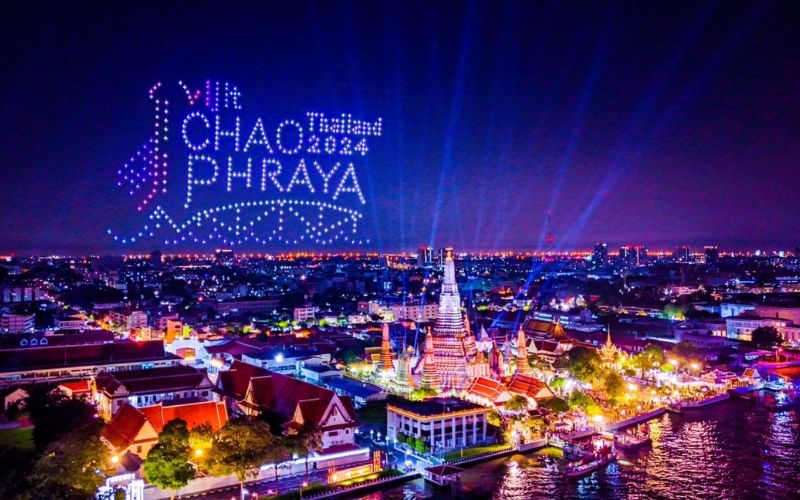 A vibrant night view of the Chao Phraya River in Bangkok with colorful lights, laser projections, and Wat Arun illuminated during an evening cultural show