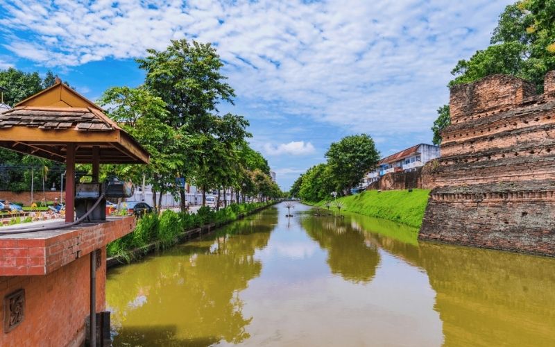 A scenic view of the wide moat surrounding the Chiang Mai Old City, showing the historic red brick walls on the right and a tree-lined street with a small pavilion on the left under a bright blue sky.