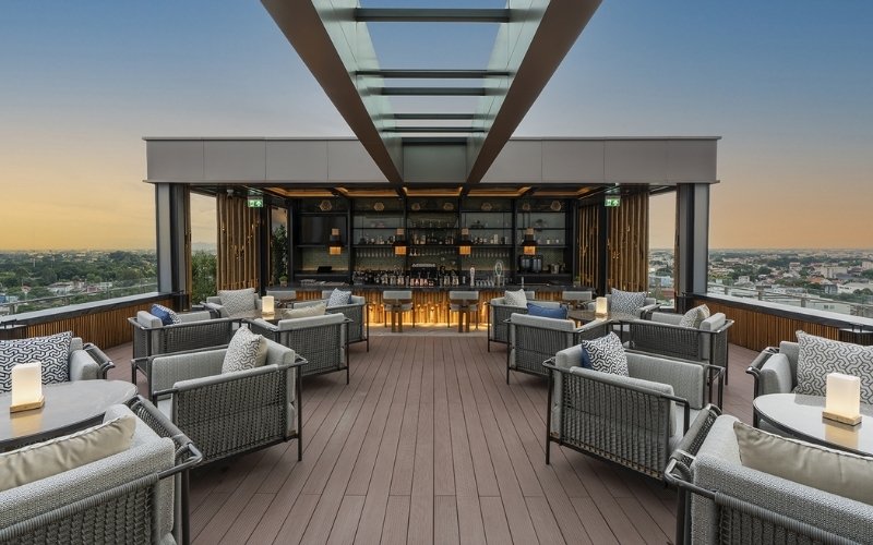 A modern, chic rooftop bar and lounge area at sunset, featuring a dark wood deck and comfortable gray cushioned seating arranged around a central bar counter. The bar has an open-air design with a glass roof structure overhead, offering panoramic views of the city skyline and surrounding landscape.
