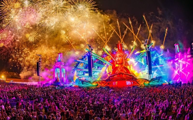 A spectacular night photo of the main stage at a massive music festival, featuring a large, brightly lit central structure shaped like a woman's face, surrounded by elaborate neon frames, lasers, and fireworks exploding above a dense crowd of concertgoers.