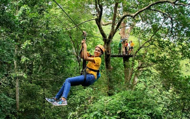 A woman smiling while ziplining through a lush green forest at Flight of the Gibbon in Chiang Mai, with other participants waiting on a wooden platform in the background.