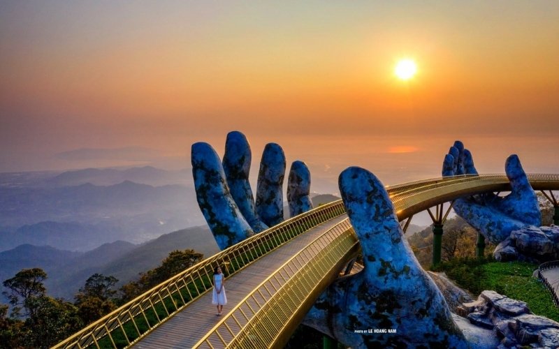A woman in a white dress walks along the Golden Bridge in Da Nang, Vietnam, held by two giant stone hands at sunrise with misty mountains in the background.