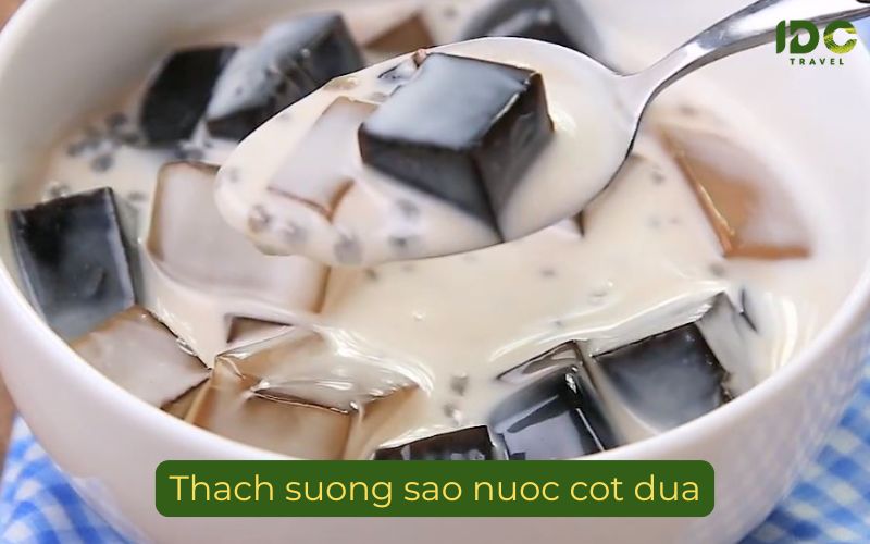 A close-up shot focusing on a metal spoon lifting a single cube of black grass jelly and some basil seeds from a bowl filled with white coconut milk and other cubes of jelly. The milk is speckled with thousands of tiny, clear basil seeds. The label "Thach suong sao nuoc cot dua" is at the bottom, and the IDC Travel logo is in the top right.