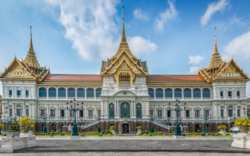 The Grand Palace in Bangkok, Thailand, featuring ornate Thai-style spires, golden roofs, and a grand courtyard under a bright blue sky.