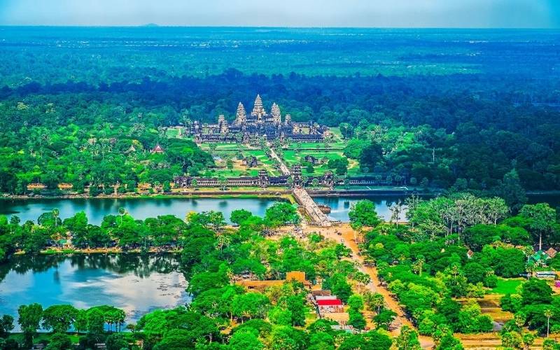Aerial view of Angkor Wat temple surrounded by lush green forests and moats in Siem Reap, Cambodia, under a clear blue sky.
