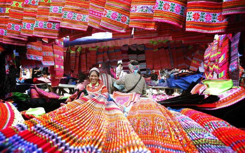Colorful highland market in Northern Vietnam, with ethnic minority women selling traditional woven fabrics under vibrant red and orange canopies