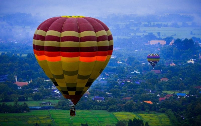 A colorful hot air balloon flying over a lush green landscape with villages and trees below, with another balloon visible in the distance under a hazy blue sky.
