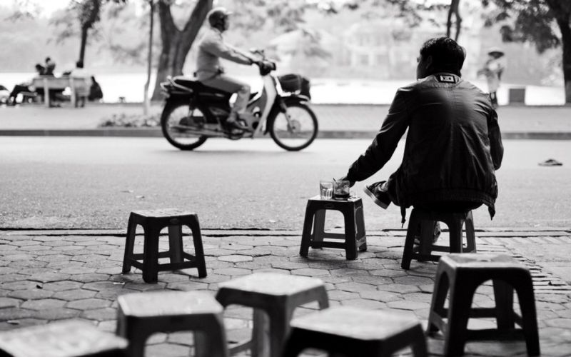 A man sits alone on a low plastic stool enjoying coffee and quietly watching the street in Hanoi, capturing the city's slow-paced coffee culture