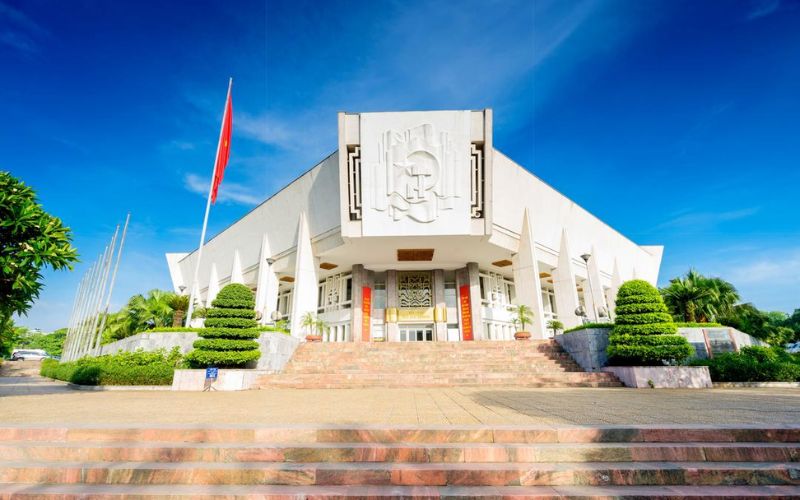 A wide-angle view of the Ho Chi Minh Museum in Hanoi, featuring its massive white concrete structure designed to resemble a blooming lotus flower, surrounded by a paved plaza and manicured trees.