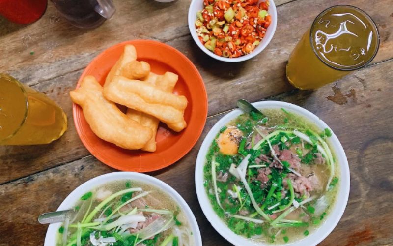 Traditional Vietnamese pho served with quẩy (fried dough), herbs, sauces, and iced tea on a rustic wooden table