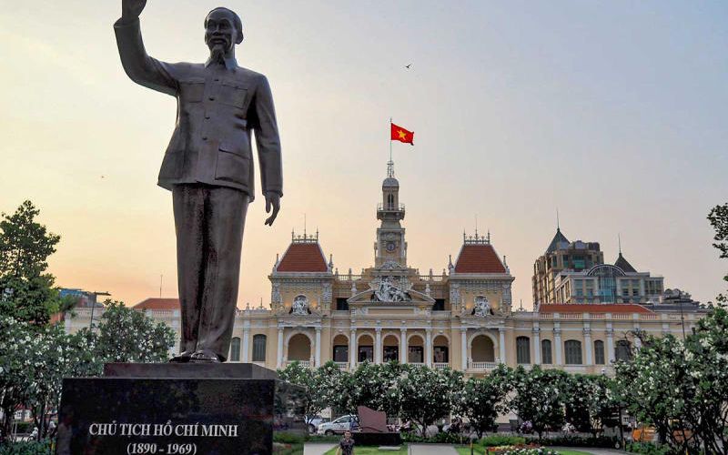 A frontal, symmetric view of the historic People's Committee Building (City Hall) on Nguyen Hue street. The grand French colonial-style facade, painted yellow with white trim, is well-lit under a bright blue sky. Several people on motorbikes and a white car are in the street in the foreground, with the Vietnamese flag flying high on the central mast.