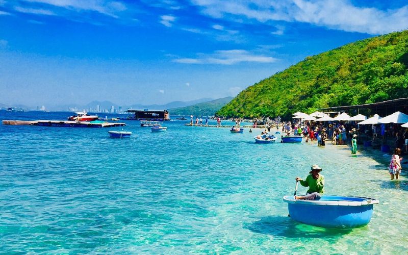 Hon Mieu Island near Nha Trang with clear blue water, visitors enjoying the beach, small round basket boats, and green hills in the background.