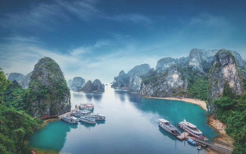 A stunning view of Ha Long Bay in Vietnam, featuring calm turquoise waters, limestone karsts, and several cruise boats anchored near small beaches surrounded by lush green cliffs.