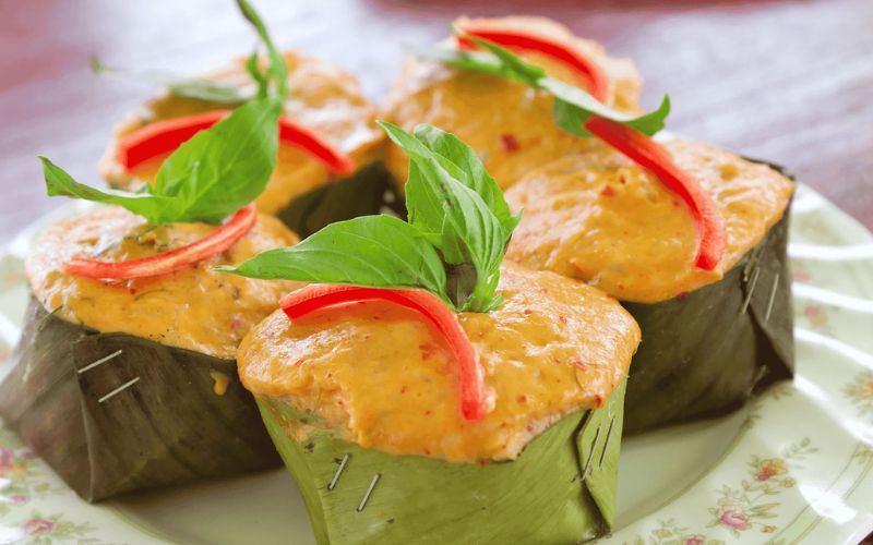 Close-up of Thai hor mok pla, steamed fish curry custard served in folded banana leaf cups, topped with creamy orange curry mixture, fresh basil leaves, and thin slices of red chili on a floral plate.