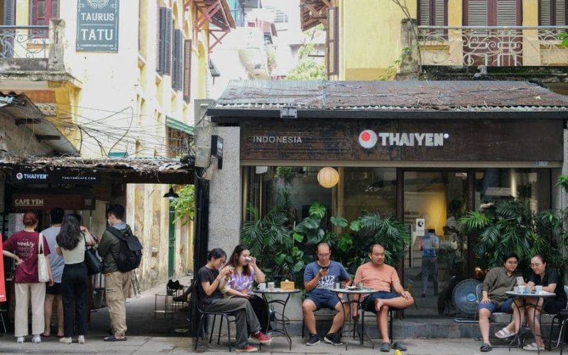 Hanoians enjoying their morning coffee on low stools outside a café, surrounded by old buildings and a relaxed street vibe