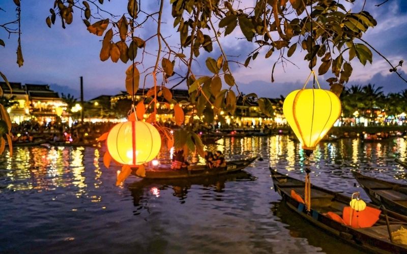 Colorful lanterns hanging from a tree illuminate the river in Hoi An, Vietnam, where boats float under the evening sky during the Full Moon Lantern Festival.