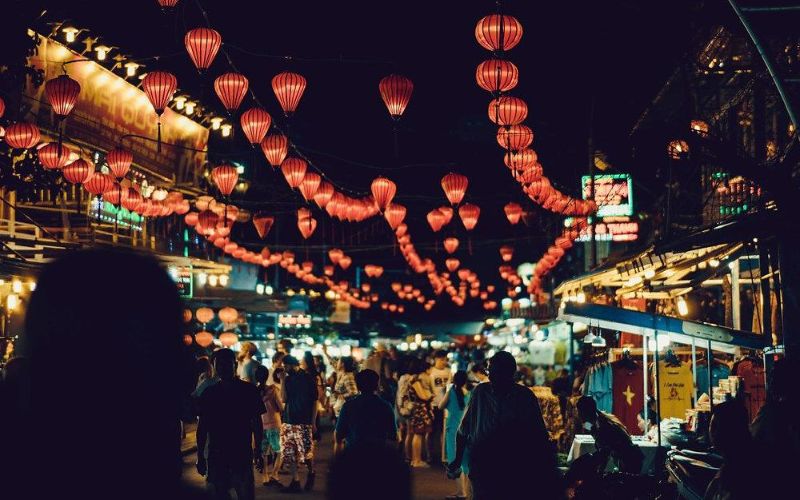 A bustling night scene at the Hanoi Old Quarter Night Market with overhead strings of glowing red lanterns and crowds of people browsing street-side stalls.