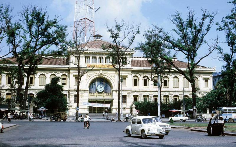 A wide exterior view of the Saigon Central Post Office in Ho Chi Minh City, showcasing its grand French colonial architecture. The building features a bright yellow facade with white decorative moldings, a prominent central clock above the arched entrance, and green-shuttered windows. In the foreground, a few tourists and local street vendors are seen on the spacious red-tiled sidewalk under a clear sky.