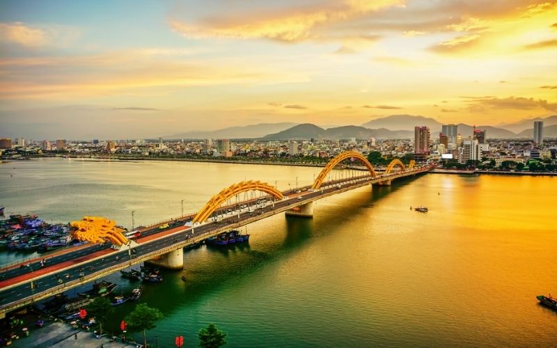 A picturesque daytime photo of the Han River in Danang, Vietnam, reflecting the warm glow of sunset. The iconic golden Dragon Bridge spans the wide river, with the city's modern skyline and mountains visible in the distance.