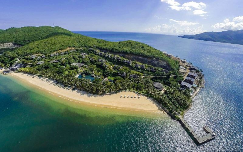Aerial view of Hon Tam Island with a curved sandy beach, turquoise water, lush green hills, and resort villas along the coastline.