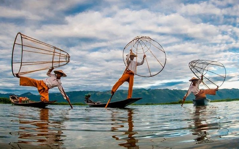 Three traditional Intha fishermen on Inle Lake, Myanmar, balancing on narrow boats while holding conical fishing nets and rowing with one leg under a cloudy sky.