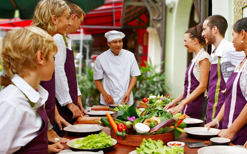Travelers of all ages gather around a local chef, ready to learn the secrets of Vietnamese cuisine using fresh herbs and colorful market ingredients.