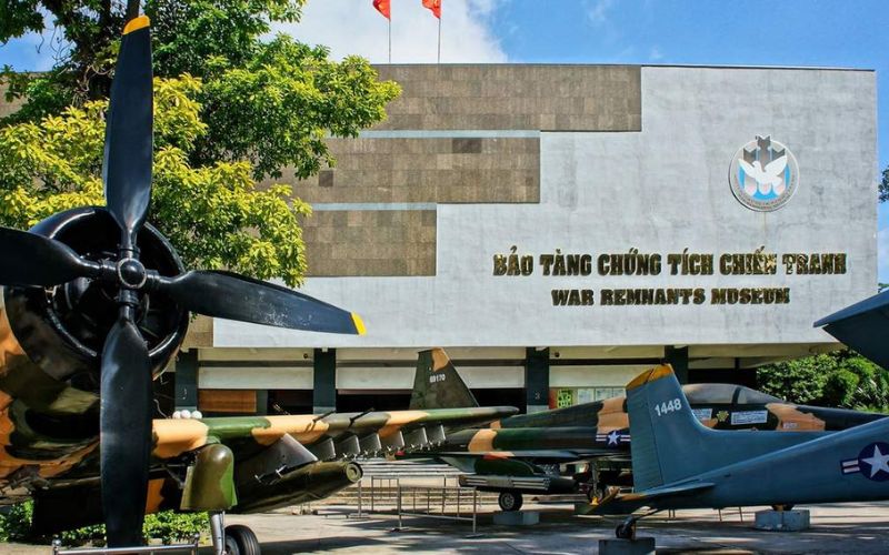 A side view of the War Remnants Museum building in Ho Chi Minh City, partially framed by the propeller of a vintage military aircraft in the foreground, with several other fighter jets parked on the museum grounds.
