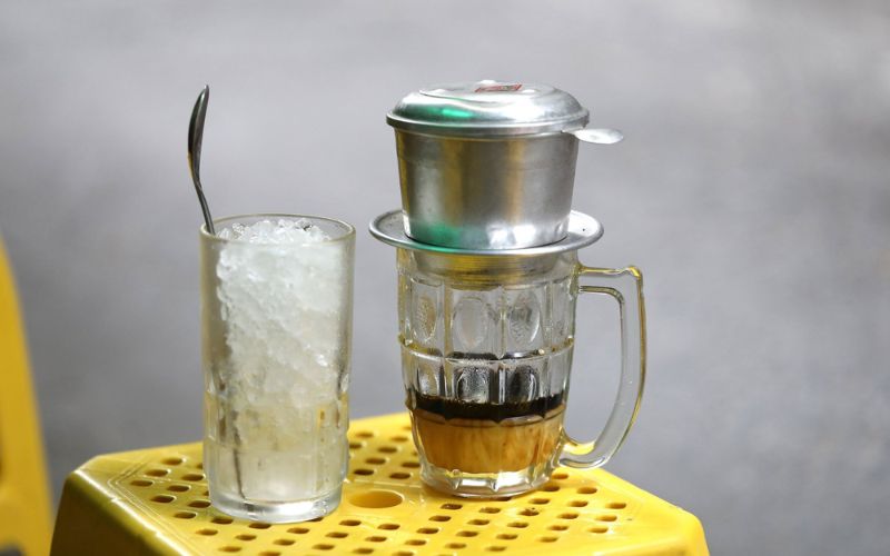 Traditional Vietnamese iced milk coffee being brewed with a phin filter, next to a glass of ice on a yellow plastic stool