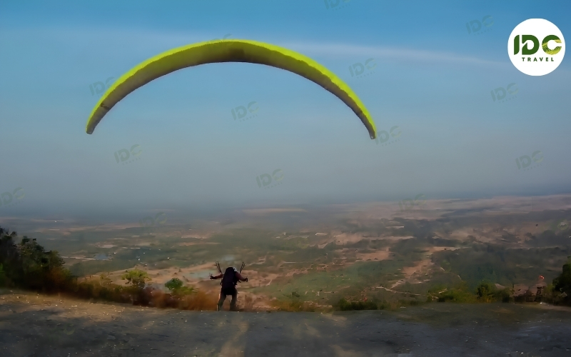 Paraglider launching with a yellow wing from a hilltop overlooking a wide valley in Thailand.