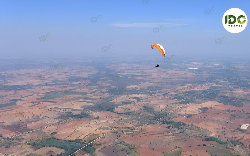 Paraglider with an orange and white wing flying high above patchwork farmland in Thailand.